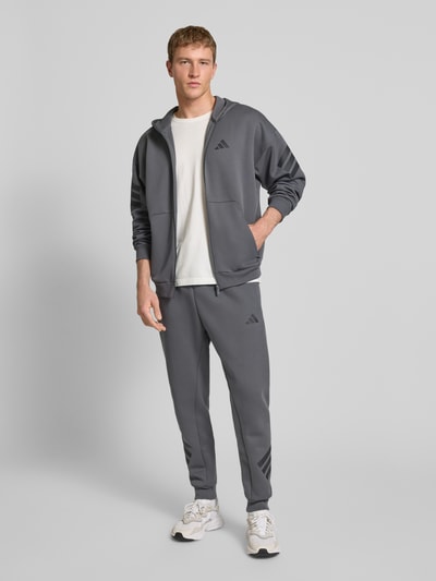 Männermodel trägt Sweatjacke mit Kapuze von adidas Sportswear in Grau mit Reißverschluss, Label-Print und Kapuze, Ganzkörperansicht von vorne.