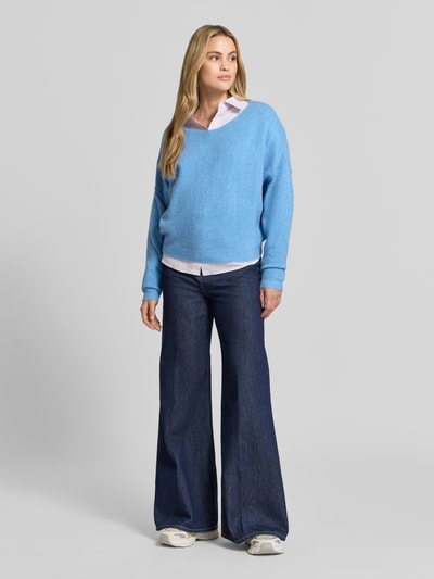 Frauenmodel trägt Relaxed Fit Strickpullover aus Material-Mix mit Leinen-Anteil von American Vintage in Hellblau über Hemd, gerippte Abschlüsse, überschnittene Schultern, Rundhalsausschnitt, langärmlig, Ganzkörperansicht von vorne.