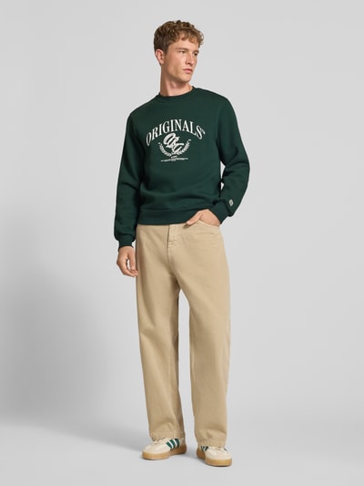 Männermodel trägt Sweatshirt mit Label-Print Modell 'FREDERIKSBERG' von JACK & JONES, dunkles Grün mit beigem Print, Rundhals, Baumwoll-Mix, Regular Fit, beigefarbene Hose, Ganzkörperansicht von vorne.