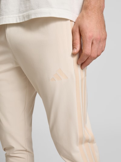 Mężczyzna nosi Jogpants mit elastischem Bund od adidas Sportswear, beżowy kolor, tkanina z domieszką stretchu, label print, detal nogawki, zbliżenie na biodro