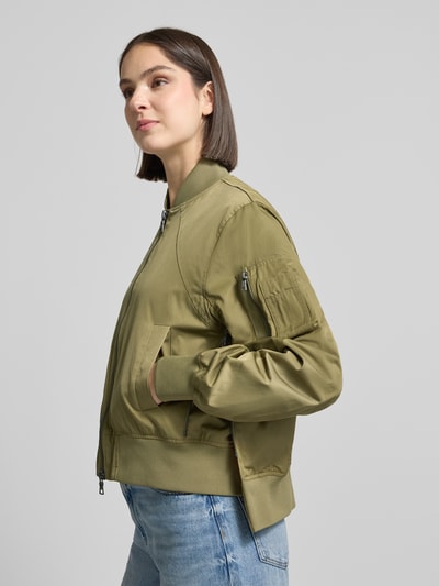 Vrouwenmodel draagt Bomberjacke mit Reißverschlusstasche am Ärmel van Blonde No. 8 in olijfgroen, materiaalmix met katoen, loose fit, opstaande kraag, tweewegritssluiting, steekzakken, elastische boorden, detailopname zijaanzicht.