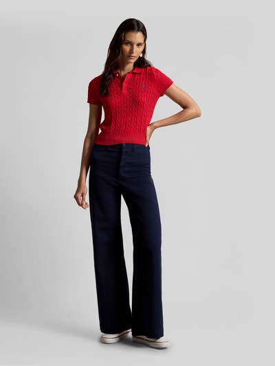 Frauenmodel trägt Regular Fit Strick-Poloshirt aus reiner Baumwolle von Polo Ralph Lauren, unifarben rot mit Zopfmuster, kurze Ärmel, Polokragen, Label Stitching, Rippenabschlüsse, kombiniert mit dunkler Hose, Ganzkörperansicht von vorne.