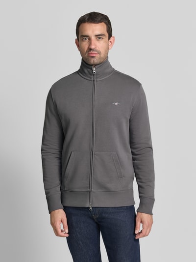 Männermodel trägt Regular Fit Sweatjacke aus Baumwoll-Mix von GANT in Grau mit Reißverschluss und Kängurutaschen, Ganzkörperansicht von vorne.