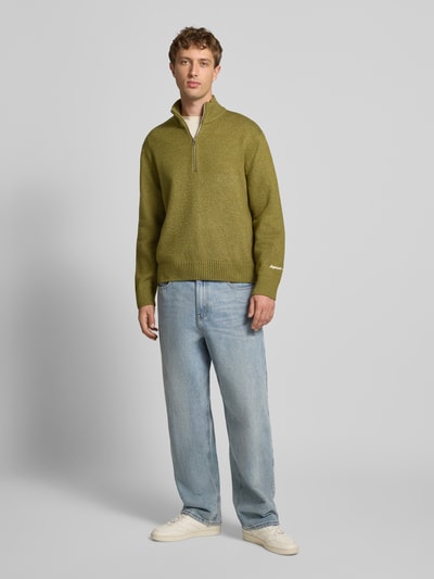Männermodel trägt Strickpullover mit gerippten Abschlüssen Modell 'NORREBRO' von Jack & Jones in Olivgrün, Ganzkörperansicht von vorne. Baumwoll-Mix mit Stretch, Regular Fit, Stehkragen, kurzem Reißverschluss und Label-Stitching.