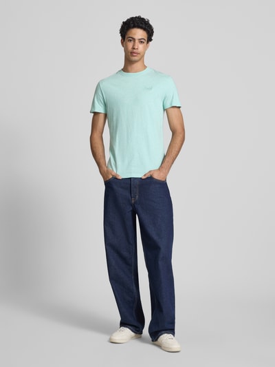 Männermodel trägt Regular Fit T-Shirt mit Logo-Stitching von Superdry in Mintgrün, kombiniert mit blauer Jeans, Ganzkörperansicht von vorne.