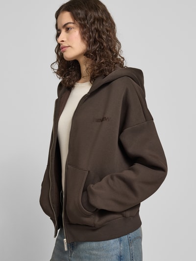 Modelka nosi Sweatjacke mit Logo und Känguru-Tasche od Review, mieszanka bawełny, krój relaxed fit, kaptur, zamek błyskawiczny, kieszeń kangurka, logo, widok z przodu do połowy ciała.