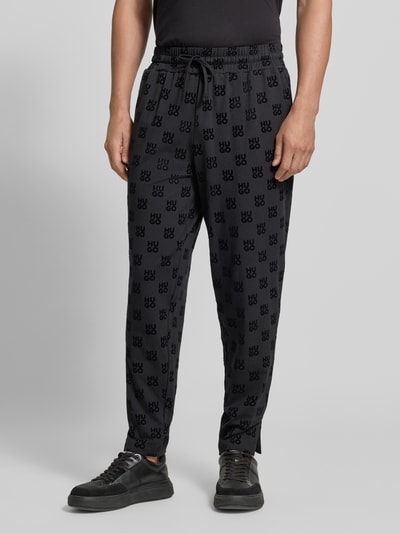 Männermodel trägt Regular Fit Sweathose aus Baumwoll-Mix von HUGO in Schwarz mit Allover-Logomuster und elastischem Bund, Nahaufnahme Unterkörper. Herren Sweathose von HUGO.