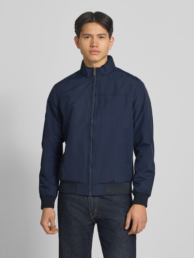 Männermodel trägt Jacke mit Stehkragen und Eingrifftaschen von Superdry in Dunkelblau, Regular-Fit, Reißverschluss, Ganzkörperansicht von vorne, Rückenlänge bei Größe M: 70 cm, Schulterbreite: 44 cm, Brustweite: 41 cm, Ärmellänge: 68 cm.