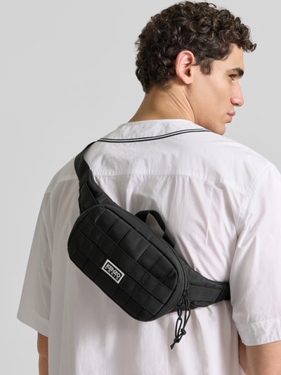 Männermodel trägt Bauchtasche mit Zweiwege-Reißverschluss von Kenzo, Label-Patch, Strukturmuster, Schwarz, Ganzkörperansicht Rückansicht. Breite 28 cm, Länge 13 cm, Tiefe 6 cm, kurzer Schulterriemen, Hauptfach