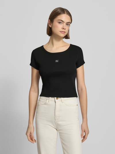 Kobiece modelka nosi Cropped T-Shirt mit Label-Detail od Calvin Klein Jeans z mieszanki bawełny i elastanu, okrągły dekolt, krótki krój, czarny, długość tyłu 43 cm dla rozmiaru S, widok całej sylwetki z przodu.