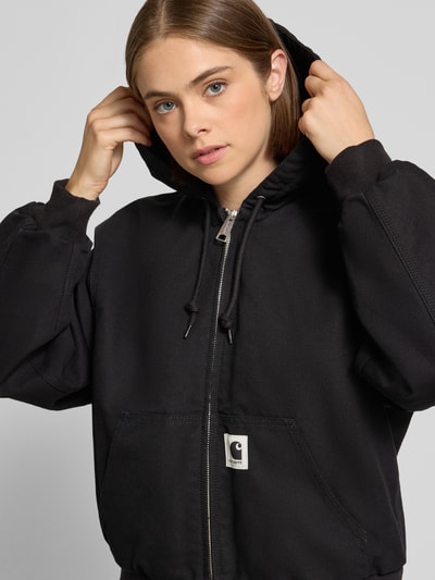 Frauenmodel trägt Bomberjacke aus reiner Baumwolle von Carhartt Work In Progress in Schwarz mit Känguru-Tasche, Nahaufnahme Oberkörper. Lockere Passform, Kapuze mit Tunnelzug, Reißverschluss, gerippte Abschlüsse, Label-Patch.