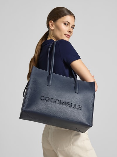 Kobiece modelka nosi Pochette mit Tragehenkeln und Logo Modell 'MYRTHA26' od Coccinelle, kolor granatowy, faktura skóry, Szerokość w rozmiarze 1: 36 cm, długość: 28 cm, głębokość: 13 cm, długość paska: 67 cm, główna komora na zamek, logo, widok całej sylwetki z tyłu.