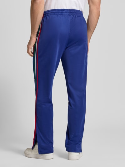 Männermodel trägt Regular Fit Sweatpants mit mehrfarbigen Kontraststreifen Modell 'FIREBIRD TP' von adidas Originals, Rückansicht Ganzkörperansicht. Unifarbenes Blau, elastischer Bund, seitliche Reißverschlusstaschen, Logo-Stitching, glatte Textur.