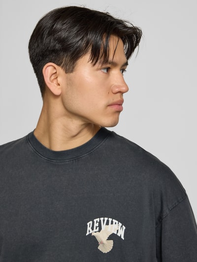Männermodel trägt T-Shirt mit Logo- und Motiv-Print von REVIEW in Schwarz, Reine Baumwolle, Loose-Fit, Gerippter Rundhalsausschnitt, Nahaufnahme Oberkörper.