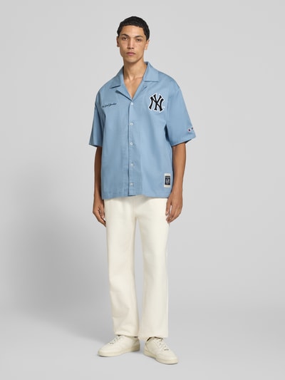 Männermodel trägt Relaxed Fit Freizeithemd aus Baumwoll-Mix von Champion in Hellblau, schwarze Statement-Stitchings, kurzer Schnitt, weiße Hose, Ganzkörperansicht von vorne.