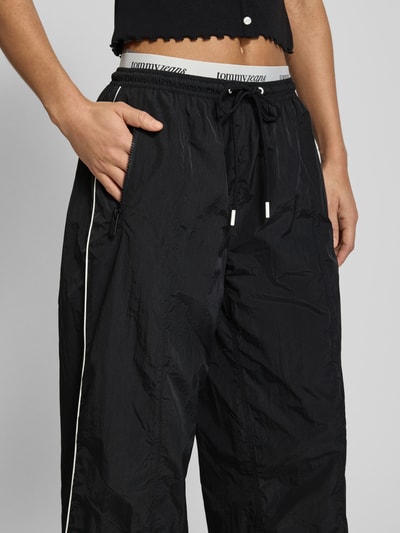 Kobiece modelka ma na sobie Regular Fit Trackpants mit Kontraststreifen od Tommy Jeans, efekt: jednolity kolor, krój regular fit, aplikacja: paski w kontrastowym kolorze, pas z logo, zapięcie: tunel, ściągacz: elastyczny, dwuwarstwowy pas, boczne kieszenie zapinane na zamek błyskawiczny, kieszeń z tyłu, kolor czarny, tekstura lekko połyskująca, widoczna struktura materiału, ujęcie: zbliżenie na dolną połowę ciała z przodu.