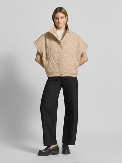 Frauenmodel trägt Cape mit Steppnähten und Eingrifftaschen von Copenhagen Muse in Beige, Relaxed-Fit, Knopfleiste, Ganzkörperansicht von vorne, Rückenlänge 66 cm, Schulterbreite 42 cm, Brustweite 25 cm.