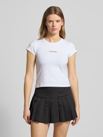 Kobiece model prezentuje T-shirt mit Rundhalsausschnitt und Label-Print von Calvin Klein Jeans, mieszanka bawełny i elastanu, krój slim fit, okrągły dekolt, nadruk z logo, długość tyłu w rozmiarze S: 42 cm, widok całej sylwetki z przodu.