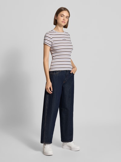 Frauenmodel trägt Barrel Fit Jeans in verkürzter Passform von GUESS, dunkelblau, 5-Pocket-Design, Logo-Detail, Ganzkörperansicht von vorne.