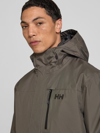 Männermodel trägt Regular Fit Jacke mit Logo-Print Modell 'Juell' von Helly Hansen in Grau, glatte Textur, Nahaufnahme Oberkörper. Herren Jacke mit Kapuze, Napoleon- und Eingrifftaschen, Rückenlänge 75 cm, Schulterbreite 48 cm, Brustweite 45 cm, Ärmellänge 70 cm.