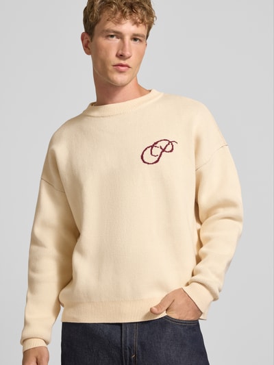 Männermodel trägt Relaxed Fit Strickpullover aus reiner Baumwolle von PROHIBITED, beige mit Label-Stitching, Nahaufnahme Oberkörper.