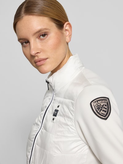 Frauenmodel trägt Steppjacke mit Label-Patches Modell 'HASKEL' von Blauer Usa in Weiß, glatte Textur, Stehkragen, Nahaufnahme Oberkörper.