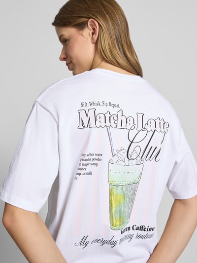 Kobieta nosi damski t-shirt z bawełny model 'LATTE' od JJXX, kolor biały, nadruk z tyłu, krój loose fit, okrągły dekolt, długość tyłu w rozmiarze S: 65 cm, widok całej sylwetki z tyłu.