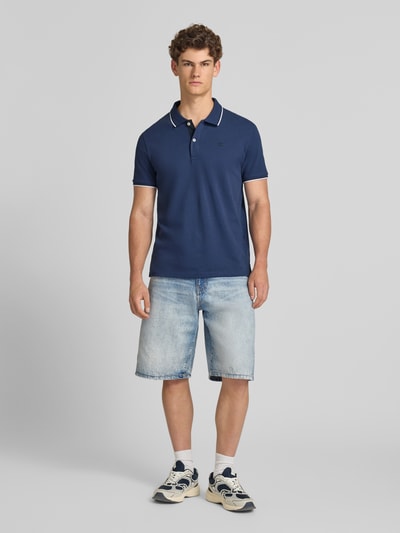 Męski model nosi Slim Fit Poloshirt mit Label-Stitching von CHAMPION z bawełny, krój slim fit, wyhaftowane logo, listwa guzikowa, długość tyłu w rozmiarze M: 73 cm, koszulka polo w kolorze granatowym, jasnoniebieskie szorty jeansowe, białe sneakersy, gładkie tło, całopostaciowy widok z przodu.