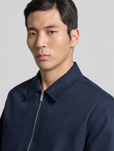 Männermodel trägt Regular Fit Blouson mit Reißverschluss Modell 'DOBBY' von CK Calvin Klein in Dunkelblau, Nahaufnahme Oberkörper. Zweiwege-Reißverschluss, Umlegekragen, Eingrifftaschen sichtbar, glatte Textur, Angaben zu Maßen im Kontext.