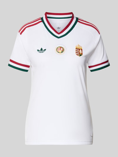 Frauenmodel trägt Ungarn Heimtrikot 2026 von adidas, Optik: Unifarben, Passform: Regular-Fit, Applikation, Logo-Stitching, Kontraststreifen, V-Ausschnitt, Ärmelform: Kurzarm, Materialverarbeitung: feine Webstruktur, Ganzkörperansicht von vorne.
