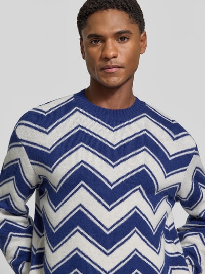 Männermodel trägt Strickpullover aus Schurwolle-Mix mit Rundhalsausschnitt von Armani Exchange, Zickzackmuster in Blau und Weiß, Regular Fit, Nahaufnahme Oberkörper. Rückenlänge und Ärmellänge bei Größe M, gerippte Abschlüsse.