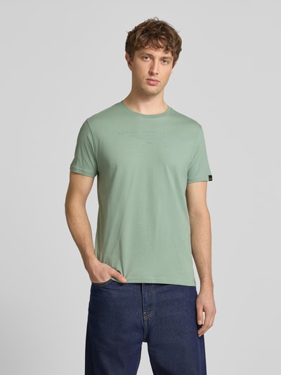 Männermodel trägt T-Shirt mit Label-Print und Rundhalsausschnitt von Alpha Industries aus Reine Baumwolle, Regular-Fit, Ganzkörperansicht von vorne. Rückenlänge bei Größe M: 67 cm.