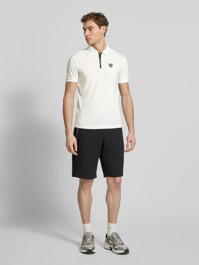 Männermodel trägt Slim Fit Poloshirt aus Viskose-Mix von EA7 Emporio Armani, weiß mit Logo-Detail, Polokragen, elastische Ärmelabschlüsse, Kombi mit schwarzen Shorts, Ganzkörperansicht von vorne.