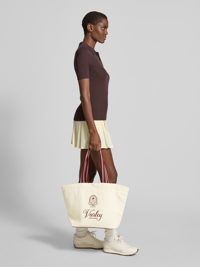 Frauenmodel trägt Shopper aus reiner Baumwolle Modell 'Robinson' von VARLEY in Creme mit Motiv-Stickereien, seitliche Ganzkörperansicht. Hauptfach mit Karabinerverschluss, Reißverschlussfach innen, Lange Tragehenkel, Label-Details.