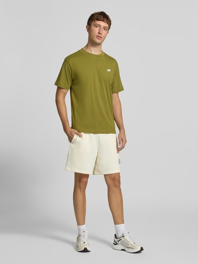 Männermodel trägt olivgrünes T-Shirt mit Label-Stitching Modell 'ESSENTIALS' von New Balance aus reiner Baumwolle, Ganzkörperansicht von vorne.