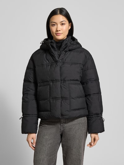 Frauenmodel trägt Cropped Fit Steppjacke mit aufgesetzten Taschen Modell 'Daisy' von Guess, Allover-Logo-Print, gefütterte Kapuze, Ganzkörperansicht von vorne.