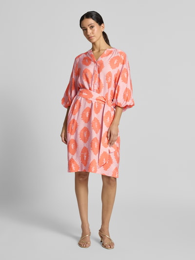 Frauenmodel trägt Knielanges Kleid aus Viskose-Mix von Christian Berg Woman in Rosa- und Orangetönen mit Taillenband und Serafino-Ausschnitt, taillierte Passform, Ganzkörperansicht von vorne.