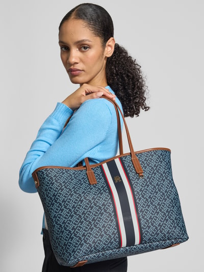 Frauenmodel trägt Shopper in Leder-Optik von Tommy Hilfiger mit Allover-Muster, Label-Applikation aus Metall und Tragehenkeln, Nahaufnahme Oberkörper.