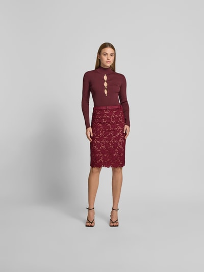 Frauenmodel trägt Slim Fit Rock mit Allover-Spitzenbesatz von Self-Portrait in Bordeaux, Knielänge, abnehmbarer Gürtel, blickdichter Unterrock, Ganzkörperansicht von vorne.