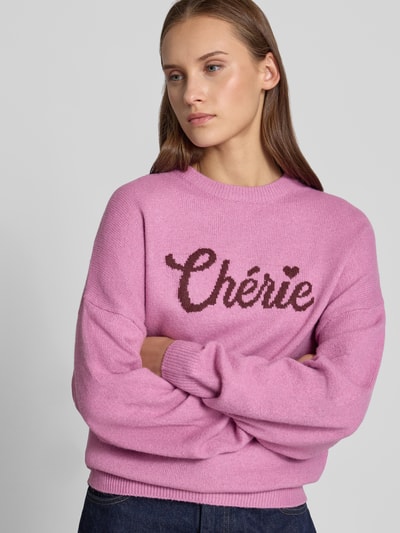 Vrouwenmodel draagt Regular Fit Pullover in strick-optik van REVIEW, materiaalmix met stretch, ribboorden, tricotlook, lange mouw, gebreide textuur, roze met donkerroze letters, bies, vooraanzicht van het bovenlichaam.