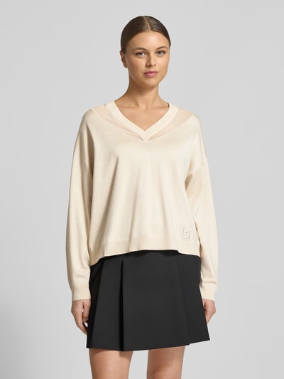 Frauenmodel trägt Relaxed Fit Pullover aus Viskose-Mix in Strick-Optik von LIU JO SPORT in Beige mit semitransparenten Partien, V-Ausschnitt, langer Ärmel, elastischen Abschlüssen, kombiniert mit schwarzem Rock; Ganzkörperansicht von vorne.