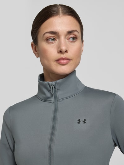 Vrouwenmodel draagt trainingsjack met opstaande kraag en logoprint van Under Armour, materiaalmix met stretch, slim fit, nahaufnahme bovenlichaam. Ruglengte maat S 59 cm, schouderbreedte 35 cm, borstwijdte 43 cm, mouwlengte 60 cm.
