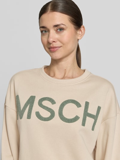 Kobieta nosi bluzę Sweatshirt mit Label-Stitching Modell 'Mela Briena' von MSCH Copenhagen, mieszanka bawełny, krój loose fit, okrągły dekolt, wyhaftowane logo, jasny beż z zielonym haftem, gładka tekstura, zbliżenie na górną część ciała.