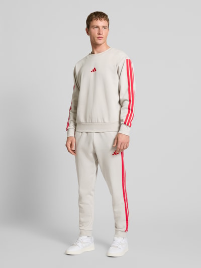 Männermodel trägt Sweatshirt mit Label-Stitching von adidas Sportswear in Beige mit roten Streifen, Baumwoll-Elasthan-Mix, Ganzkörperansicht von vorne.