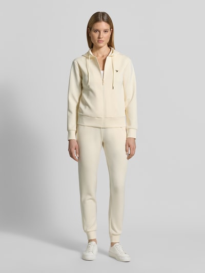 Modelka kobieca ma na sobie kremową Sweatjacke mit Label-Applikation Modell 'OLYMPE' od Guess Activewear, luźny fason, mieszanka materiałów z dodatkiem modalu, kaptur i tunel, przód ozdobiony logo, widok całej sylwetki z przodu.