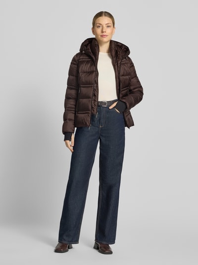 Frauenmodel trägt Steppjacke mit Kapuze und Reißverschlusstaschen von MICHAEL Michael Kors in Dunkelbraun, breite Steppnähte, gerader Schnitt, elastische Saumabschlüsse, Ganzkörperansicht von vorne.