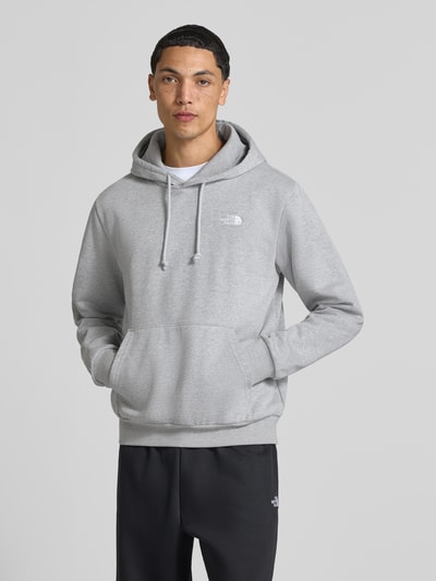 Męski model nosi bluzę z kapturem z mieszanki bawełny Model 'EVOLUTION' od The North Face, luźny krój, kieszeń kangurka, kaptur z tunelem, elastyczne wykończenia, szara, gładka tekstura, długość tyłu w rozmiarze M: 67 cm, długość rękawów w rozmiarze M: 68 cm, widok całej sylwetki z przodu.