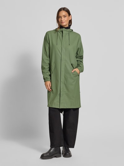 Modelka kobieca nosi Regular Fit Parker z kapturem model 'Fishtail' od Rains, zielony, kieszenie z patką, elastyczne zakończenia rękawów, krój regular fit, długość tyłu w rozmiarze S 104 cm, szerokość ramion w rozmiarze S 45 cm, szerokość w rozmiarze S 55 cm, długość rękawów w rozmiarze S 65 cm, widok całej sylwetki z przodu.