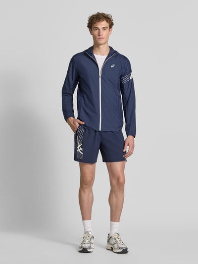Męski model nosi Regular Fit kurtkę z nadrukiem z logo Model 'ICON' od ASICS TIGER, granatowa z białym zamkiem i kapturem, krój regular fit, stójka, elastyczne wykończenia, długość tyłu w rozmiarze M: 72 cm, długość rękawów w rozmiarze M: 67 cm, widok całej sylwetki z przodu.