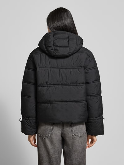 Frauenmodel trägt Cropped Fit Steppjacke mit aufgesetzten Taschen Modell 'Daisy' von Guess in Schwarz mit Allover-Logo-Print, gefütterter Kapuze, Tunnelzug an Kapuze und Ärmelabschlüssen, Rückenlänge 60 cm, Schulterbreite 45 cm, Brustweite 58 cm, Ärmellänge 60 cm, Ganzkörperansicht Rückansicht.